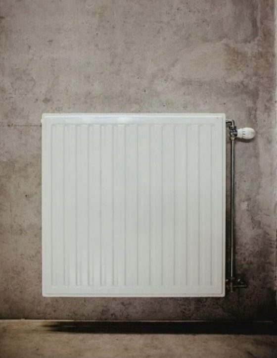 Weißer Plattenheizkörper an einer rohen Betonwand mit sichtbarem Thermostatventil und Heizungsrohr – Symbol für Wärmeversorgung in Gebäuden.